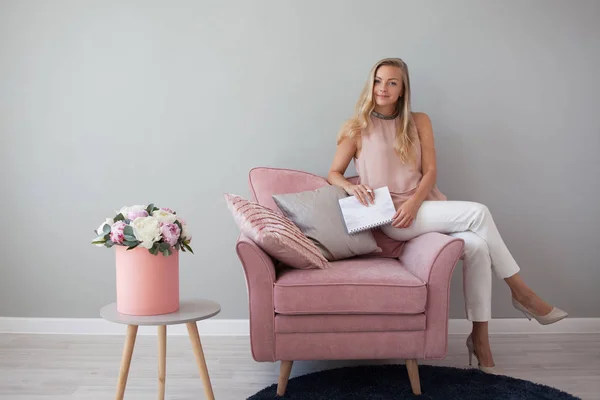 Young friendly woman sitting in a chair with a notebook in her hands. Stylish interior design, bouquet of flowers. — Stock Photo, Image