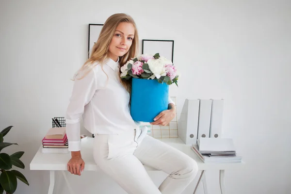 Elegante joven rubia en oficina, camisa blanca y pantalones. Sostiene un lujoso ramo de peonías en una caja de sombrero azul . —  Fotos de Stock