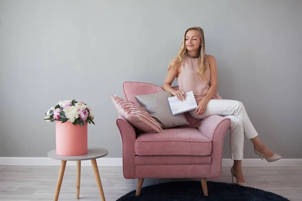 Young friendly woman sitting in a chair with a notebook in her hands. Stylish interior design, bouquet of flowers. — Stock Photo, Image