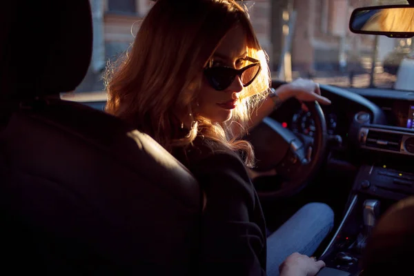 Hermosa joven con gafas de sol sentada detrás del volante de un coche. Foto del asiento trasero —  Fotos de Stock