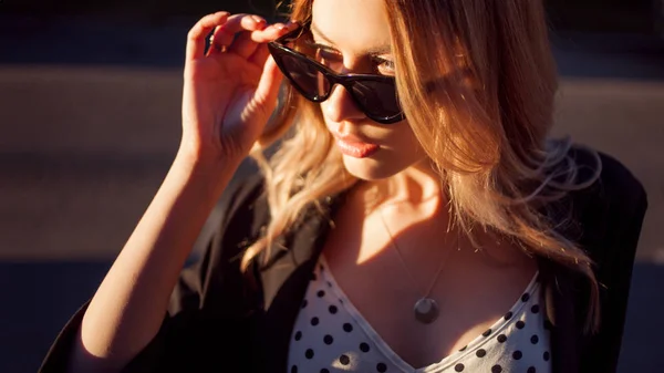 Young attractive blonde on hot summer evening Corrects sunglasses, portrait on the city background. — Stock Photo, Image