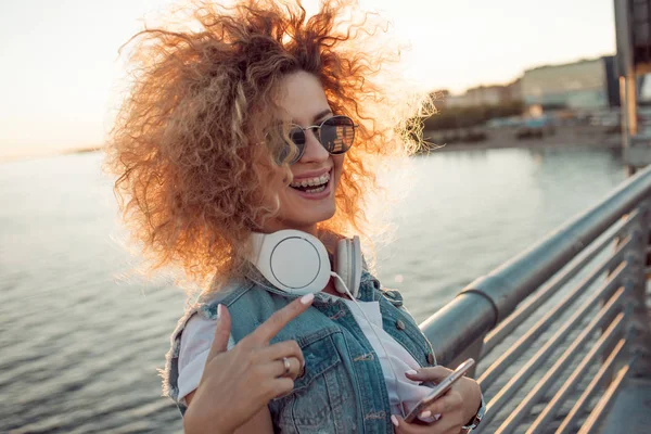 Chica de moda con auriculares grandes y gafas de sol en un paseo por la ciudad, mujer joven utiliza un teléfono inteligente —  Fotos de Stock