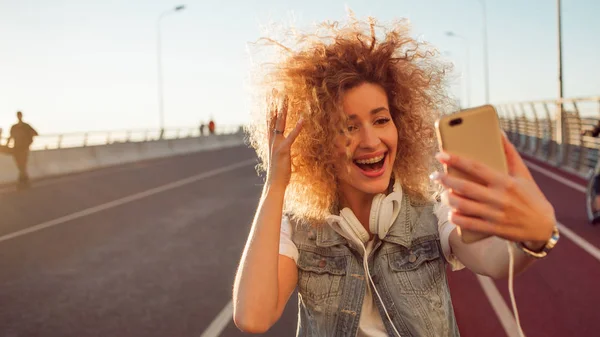 Mutlu genç kızı sokakta Smartphone'da fotoğraf al. Akıllı telefon ve kulaklık ile güzel sarışın bir selfie yapmak — Stok fotoğraf