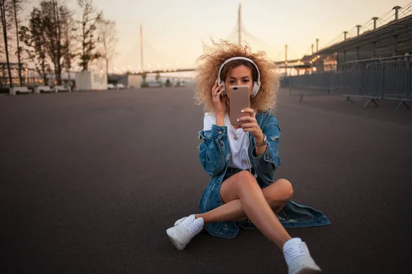 Chica joven feliz en la calle tomar una foto en el teléfono inteligente. Hermosa rubia con auriculares y smartphone hacen una selfie —  Fotos de Stock
