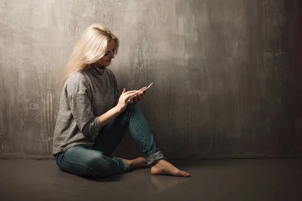 Een jonge vrouw maakt gebruik van een smartphone, zittend op de vloer en kijkend naar het scherm van de smartphone, het gebruik van het Internet — Stockfoto