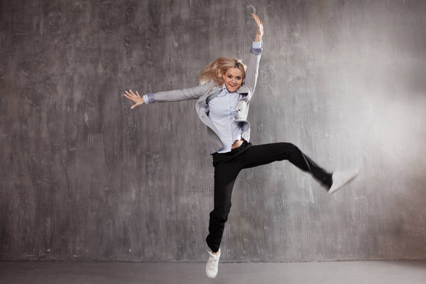 Young blonde woman in business suit and sneakers jumping for joy, gray textured background