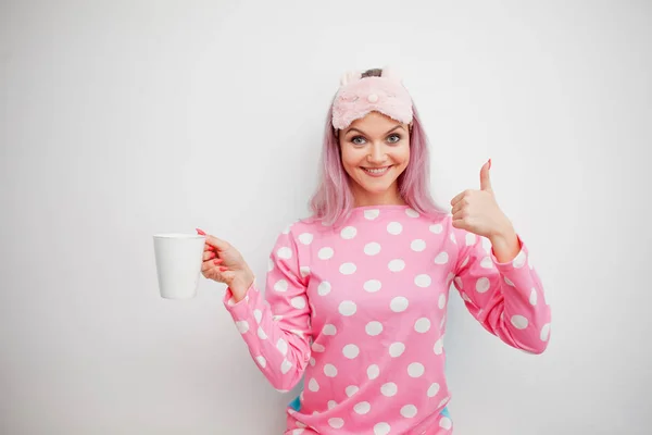 Goedemorgen. Lachende jonge vrouw duim verschijnt en drinkt koffie. Meisje in roze pyjama en slaap masker — Stockfoto