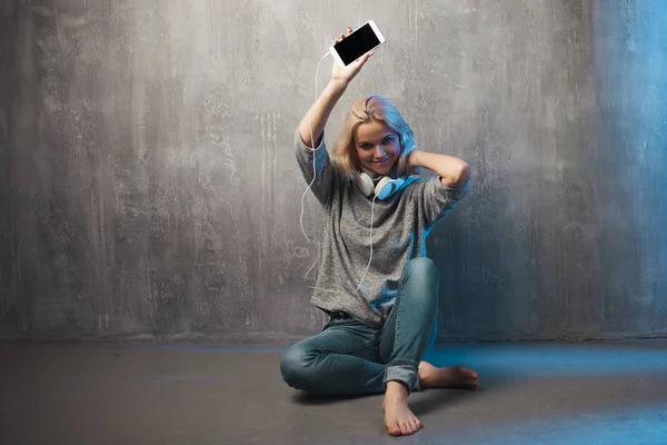 Mujer joven y atractiva con smartphone y auriculares grandes, sentada en el suelo. Música en la aplicación móvil . — Foto de Stock