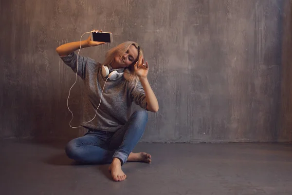 年轻和有魅力的女人与智能手机和大耳机, 坐在地板上。移动应用程序中的音乐. — 图库照片