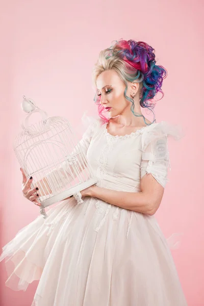 Menina elegante em vestido branco. Coloração de cabelo criativo. Penteado multicolorido. Conceito com uma gaiola de pássaro vazia . — Fotografia de Stock