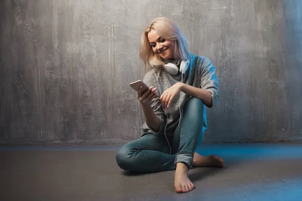 Mujer joven y atractiva con smartphone y auriculares grandes, sentada en el suelo. Música en la aplicación móvil . —  Fotos de Stock