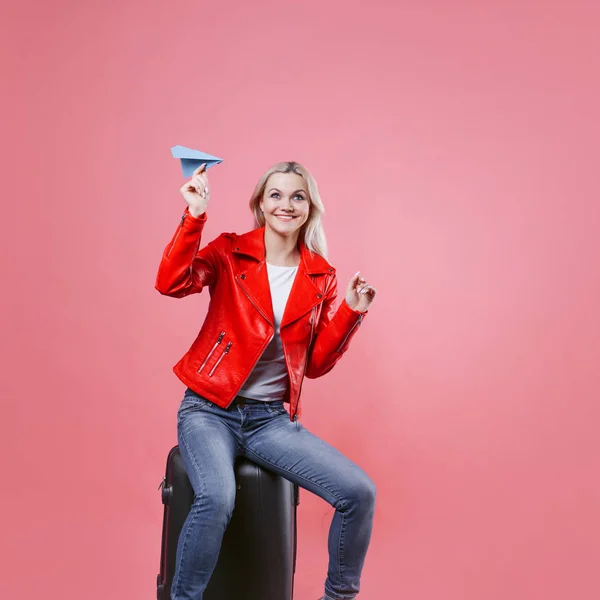 Gelukkig jonge vrouw met reizen koffer lanceert papieren vliegtuigje. Blonde toeristische meisje op roze achtergrond, concept — Stockfoto