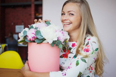 Bir şapka kutusunda çiçekli mutlu sarışın kadın modern bir ofiste. Peonies buket.