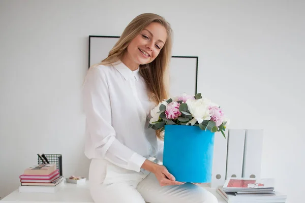 Stylish young blonde in office, white shirt and pants. Holds a luxurious bouquet of peonies in a blue hat box. — Stock Photo, Image
