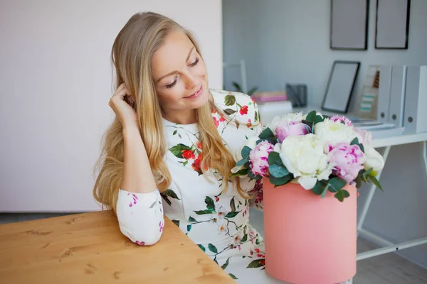 Gelukkig blonde vrouw in een modern kantoor met bloemen in een doos van de hoed. Boeket van pioenrozen. — Stockfoto