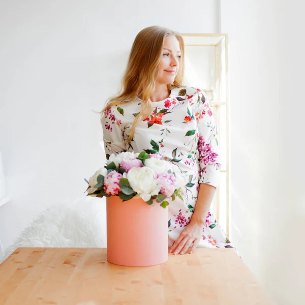 Mujer rubia feliz en una oficina moderna con flores en una caja de sombreros. Ramo de peonías . —  Fotos de Stock