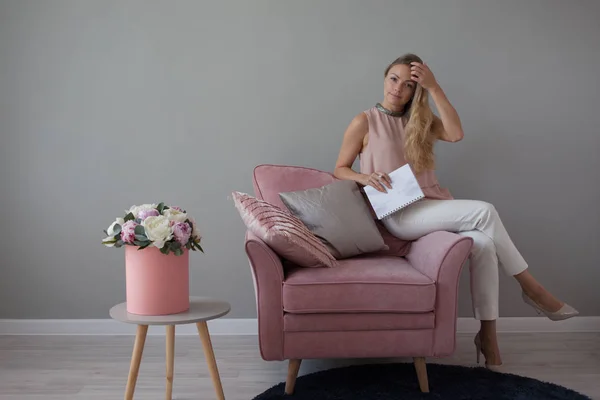 Young friendly woman sitting in a chair with a notebook in her hands. Stylish interior design, a bouquet of flowers. — Stock Photo, Image