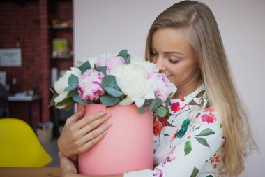 Bir şapka kutusunda çiçekli mutlu sarışın kadın modern bir ofiste. Peonies buket.