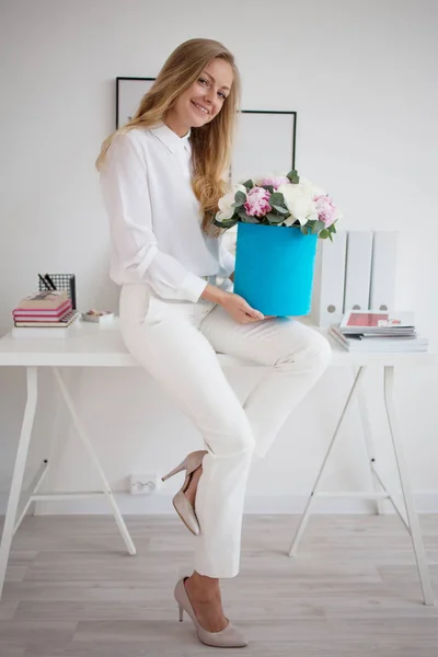 Elegante joven rubia en oficina, camisa blanca y pantalones. Sostiene un lujoso ramo de peonías en una caja de sombrero azul . — Foto de Stock