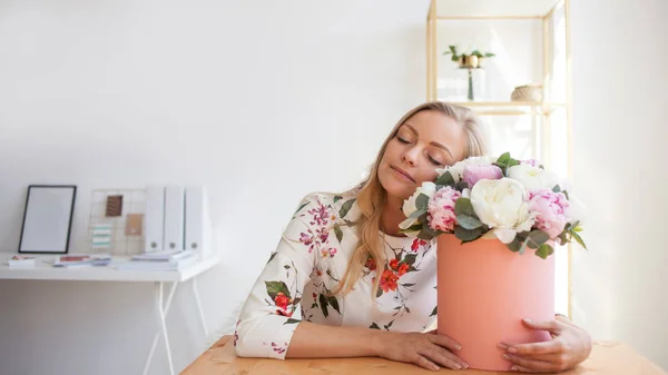 Mujer rubia feliz en una oficina moderna con flores en una caja de sombreros. Ramo de peonías . — Foto de Stock