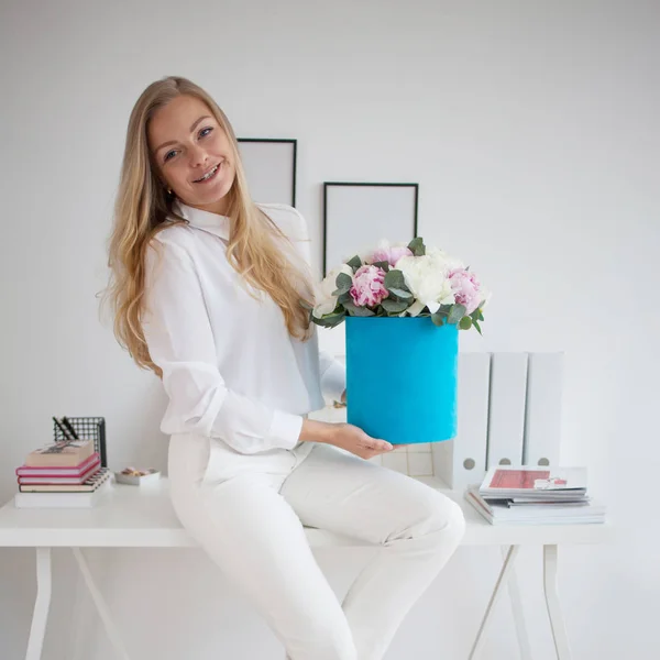 Stylish young blonde in office, white shirt and pants. Holds a luxurious bouquet of peonies in a blue hat box. — Stock Photo, Image