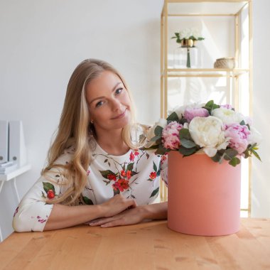 Bir şapka kutusunda çiçekli mutlu sarışın kadın modern bir ofiste. Peonies buket.