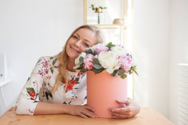 Bir şapka kutusunda çiçekli mutlu sarışın kadın modern bir ofiste. Peonies buket.