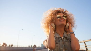 Sevimli Kıvırcık sarışın müzik dinleyin. Genç kadın büyük kulaklıklar ile. trendy kız açık havada portresi