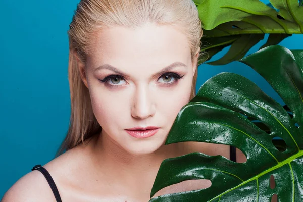 Face of attractive young blonde and leaves of palm trees. Young woman in blue bikini on blue background. — Stock Photo, Image