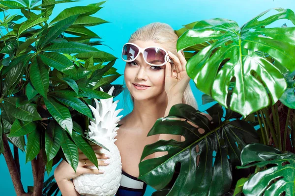 Mladá blond žena v modrých bikinách na modrém pozadí, sluneční brýle a bílé ananas — Stock fotografie