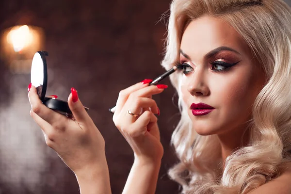Beautiful young blonde makes a bright holiday makeup. Glamorous make-up. attractive young woman paints the eyelashes — Stock Photo, Image