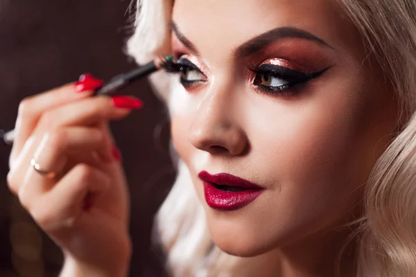Beautiful young blonde makes a bright holiday makeup. Glamorous make-up. An attractive young woman paints the eyelashes