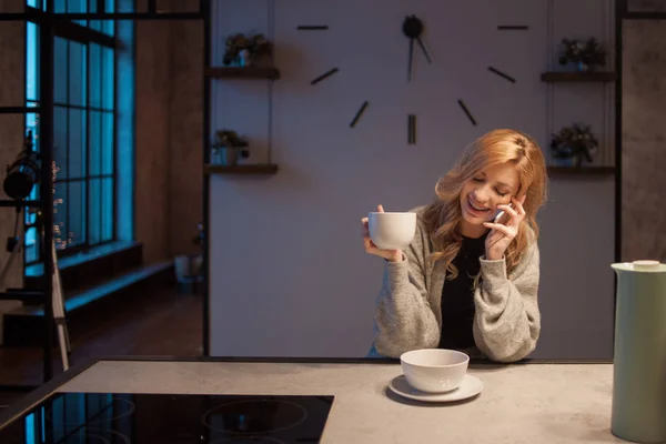 Charmerende pige i køkkenet om morgenen. Taler i telefon og spiser morgenmad. En ung kvinde med krus i hænderne - Stock-foto