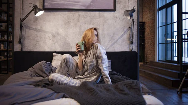 Viehättävä Nuori nainen pyjamassa, istuu sängyssä ja juo aamukahvia — kuvapankkivalokuva