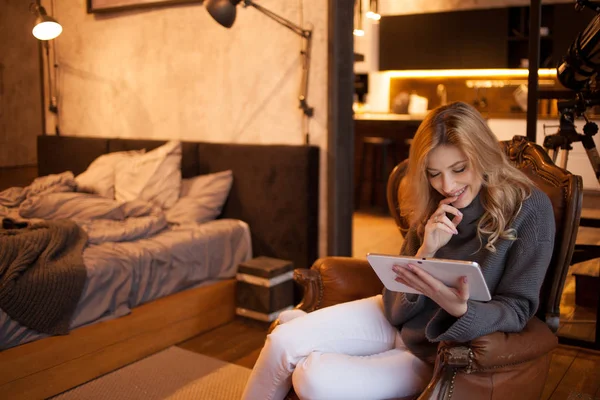 Mooi en vol vertrouwen jonge blonde zit in een comfortabele stoel in haar appartement. — Stockfoto