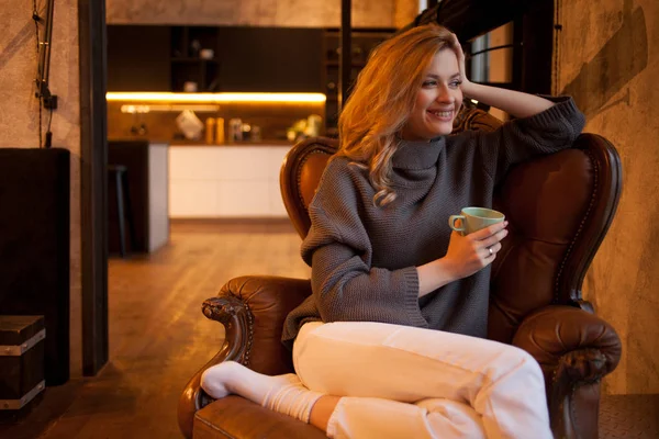 Gelukkig en lachende jonge blonde zit in een comfortabele stoel in haar appartement. — Stockfoto
