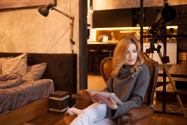 Belle et confiante jeune blonde est assise dans une chaise confortable dans son appartement . — Photo