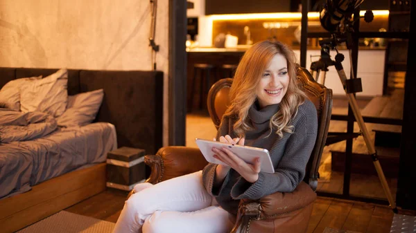 Mooi en vol vertrouwen jonge blonde zit in een comfortabele stoel in haar appartement. — Stockfoto