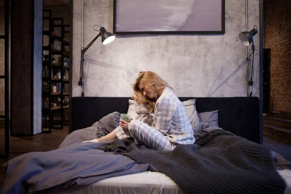 Очаровательная молодая женщина в пижаме, сидит в постели и пьет утренний кофе — стоковое фото