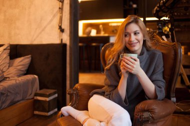 güzel ve kendine güvenen genç sarışın onun evinde rahat bir sandalyede oturuyor.