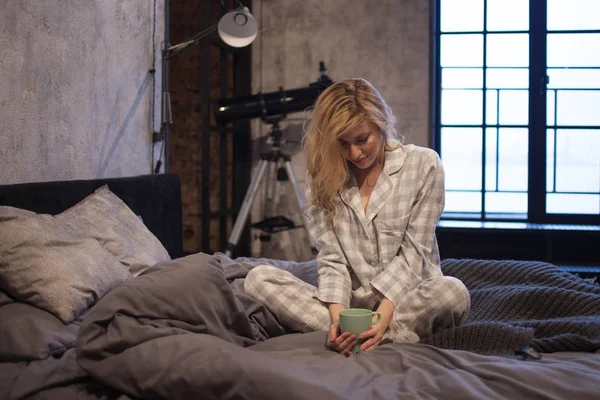 Charmante jongedame in pyjama, zittend op bed en's ochtends koffie drinken — Stockfoto