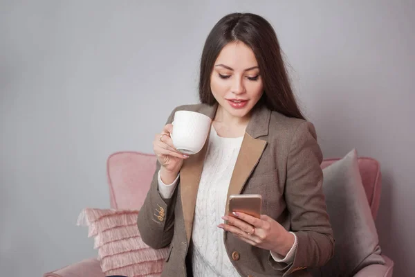 Okouzlující brunetka podnikání v bunda sedí v křesle a chatování ve smartphonu — Stock fotografie