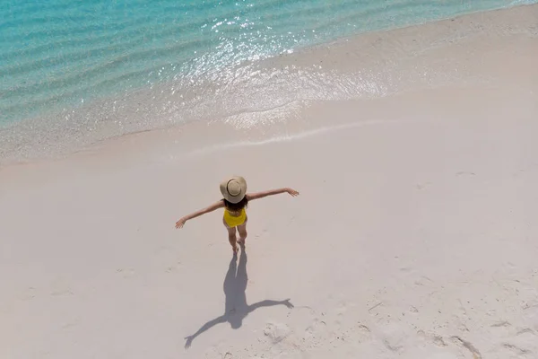 Girl alone on seashore and enjoys the sun. Young woman in a straw hat and yellow swimsuit stands with arms outstretched — Stock Photo, Image