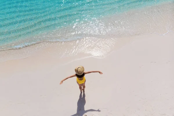 Дівчина одна на березі моря і насолоджується сонцем. Молода жінка в солом'яному капелюсі і жовтий купальник стоїть з розтягнутими руками — стокове фото
