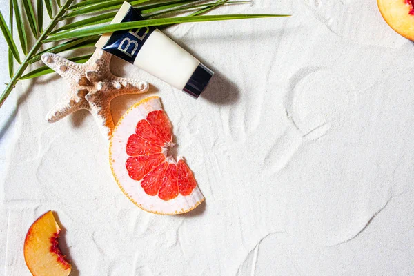 Sole sicuro. Cura della pelle. Un tubetto di crema si trova sulla sabbia della spiaggia, circondato da frutti vivaci, coralli e foglie di palma . — Foto Stock