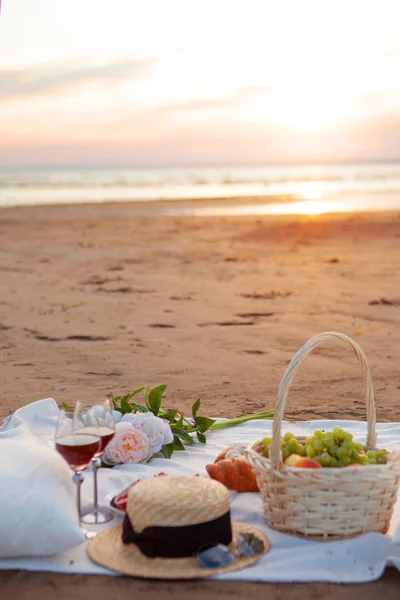 Picnic sotto i raggi del sole al tramonto sulla sabbia. Sul plaid giace cesto di frutta, bicchieri, fiori e dolci . — Foto Stock