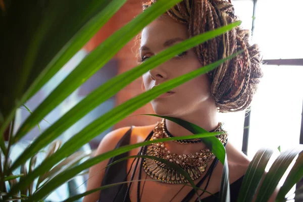 Tropical flavor. Close-up portrait of beautiful, woman with large jewels and afro braid, surrounded by tropical plants. — Stock Photo, Image