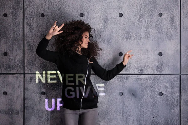 Cheer up. Cheerful young curly woman dancing on concrete wall background. Multi-colored projection says: Never give up.