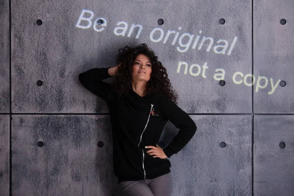 Femme bouclée posant activement sur fond de mur en béton. L'inscription illuminée colorée est projetée sur le mur . — Photo