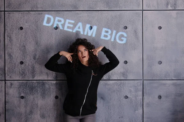 Mujer rizada posando activamente sobre fondo de pared de hormigón. Una inscripción iluminada de color se proyecta en la pared . — Foto de Stock